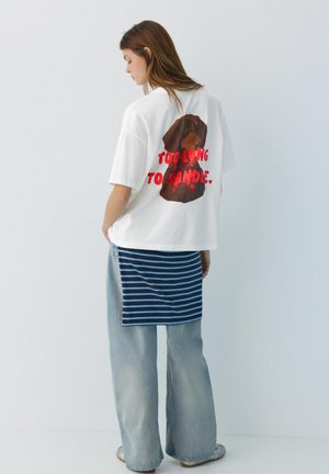 Donna con t-shirt bianca oversize con cane marrone e testo rosso "Too Long To Handle" sul retro, abbinata a una gonna a righe sopra jeans a gamba larga.