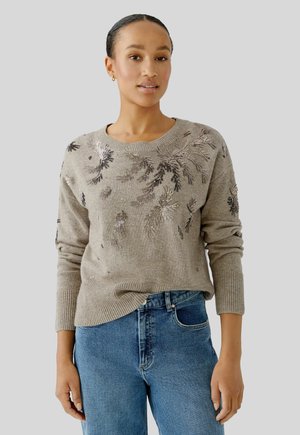 Strickpullover in Taupe mit floraler metallic Stickerei. Verfügt über einen runden Ausschnitt und gerippte Bündchen, kombiniert mit blauen Jeans.