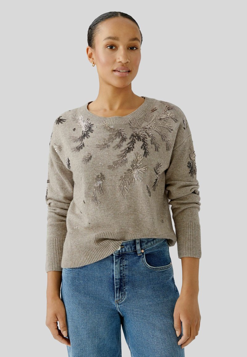 Garen gebreide trui in taupe met bloemmotieven in metallic borduursel. Heeft een ronde halslijn en geribbelde manchetten, gecombineerd met blauwe spijkerbroeken.