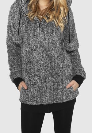 Kobieta ubrana w szarą, puszystą bluzę rozpinaną na zamek z kapturem i czarnymi mankietami, z rękami w przednich kieszeniach, w połączeniu z czarnymi spodniami.