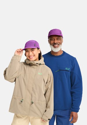 Lächelnder Mann und Frau, die violette Mützen und Outdoor-Jacken tragen, stehen nebeneinander vor einem weißen Hintergrund.