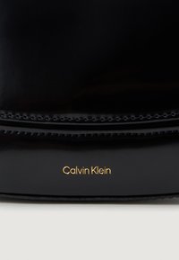 Mustan läikiva naha käekott, millel on sile pind, kuldne "Calvin Klein" logo ja puhtad jooned koos kerge õmbluse aktsendiga.