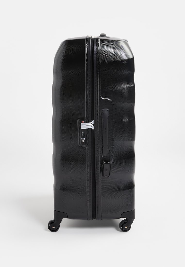 Schwarzer Hartschalenkoffer mit strukturierter Oberfläche, geriffeltem Design, seitlichem Griff, Schlossmechanismus und vier Rollen für Mobilität.