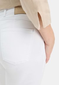 Jeans in denim bianco con un design aderente, dotati di una tasca posteriore e un dettaglio di bottone argentato. Il tessuto ha una consistenza liscia.
