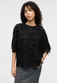 Haut à franges noires avec manches courtes, mettant en valeur un matériau texturé et superposé. Associé à une jupe grise simple, ajoutant un contraste à l'ensemble.