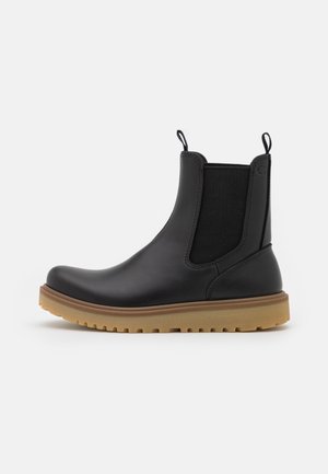 Bota Chelsea de cuero negro con suela de goma marrón claro, paneles laterales elásticos y tiradores en la lengüeta y el talón, mostrada de perfil.
