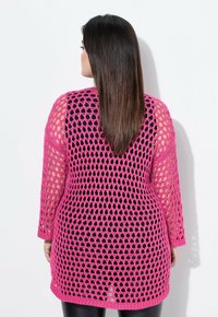 Rosa gehäkelte Strickjacke mit einem lockeren, offenen Maschenmuster, ausgestattet mit langen Ärmeln und einer längeren Passform, getragen über einem schwarzen Oberteil.