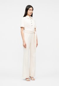 Mujer con cabello largo y negro, vestida con una blusa de manga corta color crema con botones, pantalones anchos a juego con cinturón y tacones abiertos, sobre fondo blanco.