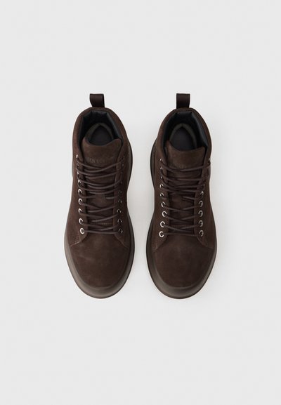 Adidași înalți din suede maro, cu ochiuri metalice, șireturi maro și căptușeală moale neagră; având un lanț de prindere la spate.