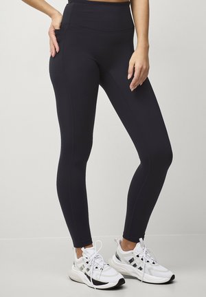 Kvinde iført sorte højttaljede leggings med sidelomme og hvide sportssko, stående mod en ensfarvet lys baggrund.