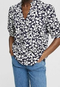 Blouse bleu marine à motif floral blanc, manches courtes bouffantes et encolure en V, en tissu léger, associée à un jean bleu en denim.