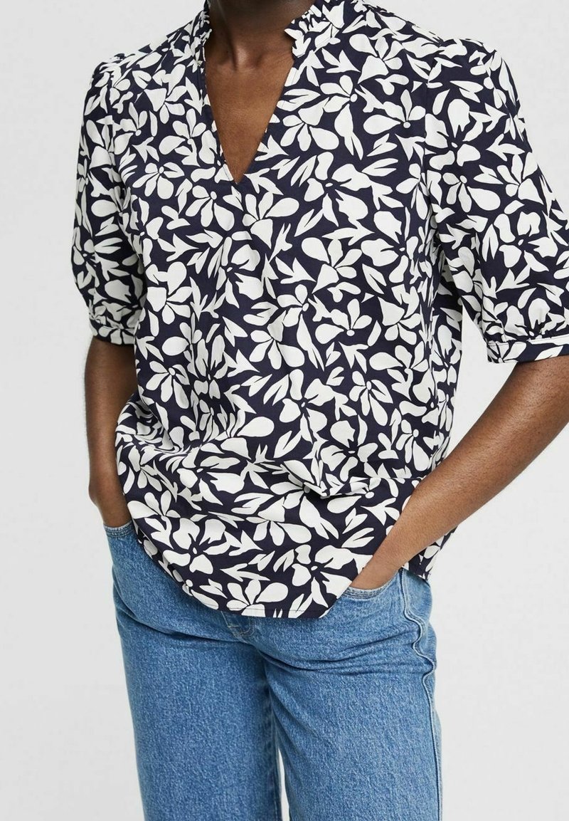 Blouse bleu marine à motif floral blanc, manches courtes bouffantes et encolure en V, en tissu léger, associée à un jean bleu en denim.