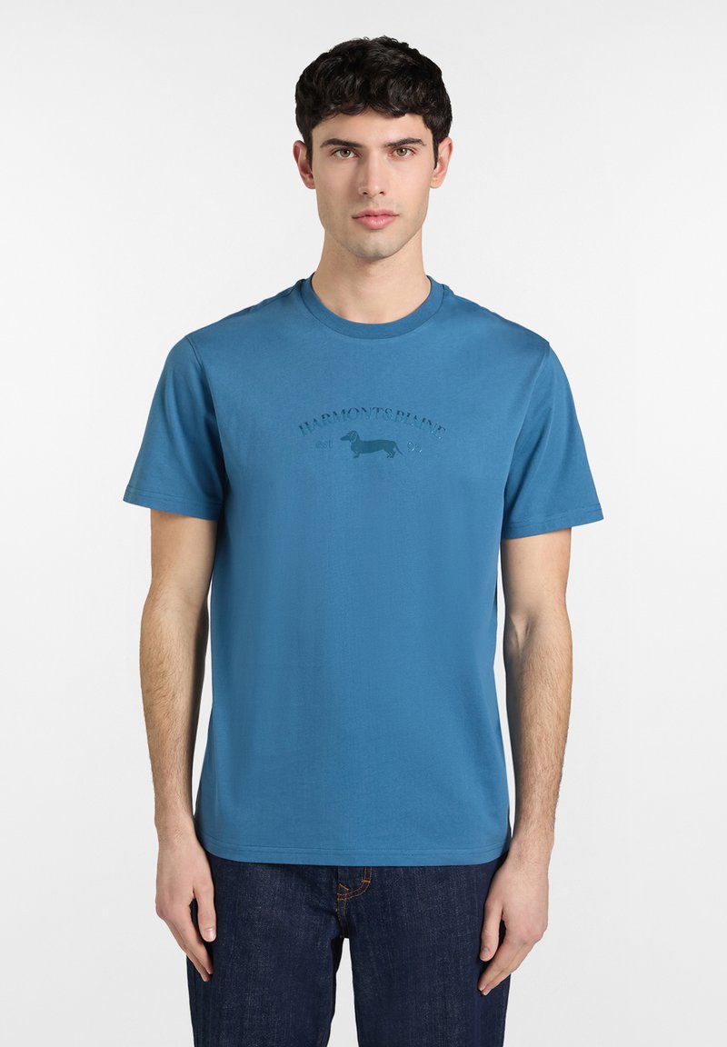 Joven con camiseta azul de manga corta con un pequeño logo de perro salchicha y texto en el pecho, combinado con vaqueros oscuros.