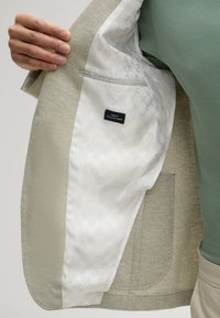 Helles graues Blazer aus einem strukturierten Gewebe. Das Innere ist mit einem glatten weißen Futter ausgestattet, das ein dezentes Muster aufweist, sowie einem Etikett mit der Aufschrift "Finest Italian Fabric."