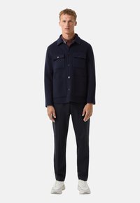 Granatowa kurtka z mieszanki wełny z kołnierzem, dwiema przednimi kieszeniami naszytymi i zapięciem na guziki. Para dopasowanych czarnych spodni i białe sneakersy.