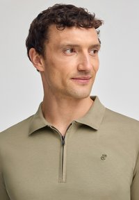 Olivenfärgad zip-neck polo skjorta, tillverkad av mjukt tyg, med en kontrasterande dragkedja och liten logotyp på bröstet. Slät textur.