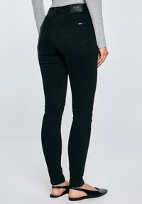 Sieviete ar slaidām melnām džinsa biksēm un melniem slingbeka apaviem, stāvot viena roka pie gurna pret vienveidīgu fonu.