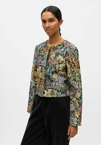 Veste à motif floral avec des manches longues, un col rond et une fermeture à l'avant. Les couleurs incluent le vert, le jaune, le violet et le marron, avec un tissu texturé.