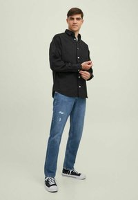 Camisa negra de botones con mangas largas, combinada con jeans de mezclilla claros desgastados y zapatillas negras con suelas blancas.