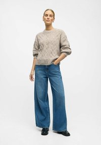 Suéter beige texturizado con un patrón de diamantes, combinado con pantalones vaqueros anchos de mezclilla azul y botas negras hasta el tobillo. El modelo está de pie con las manos en los bolsillos.