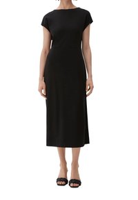 Black midi dress with cap sleeves, smooth fabric, straight cut, and a subtle slit on the side. Worn with black braided slide sandals.