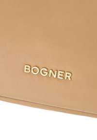 Dettaglio di un tessuto beige con lettere metalliche dorate che formano la parola "BOGNER" su una superficie testurizzata, che mostra parte di una cucitura.