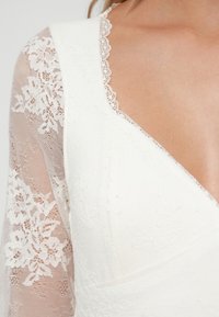 Biele šaty s kvetinovými čipkovými rukávmi, hlbokým výstrihom do V a textúrovaným telom. Jemná čipkovaná obruba okolo výstrihu pridáva detail.