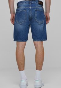 Pantalones cortos de mezclilla en un azul medio con un lavado desvaído, con un corte clásico, cinco bolsillos y una etiqueta de marca visible en la cinturilla.