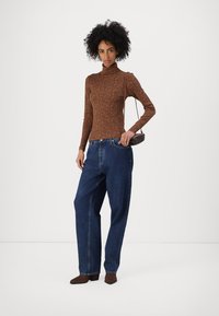 Braun gemusterter Rollkragenpullover, hoch-taillierte blaue Jeans und braune spitz zulaufende Schuhe. Schlichte Umhängetasche und strukturierte lockige Haare.