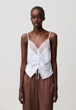 Jeune femme portant un caraco blanc orné de dentelle et un pantalon marron ample, debout devant un fond uni gris clair.