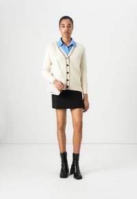 Cream knit cardigan with dark buttons, paired with a light blue collared shirt and black mini skirt. Black ankle boots complete the look.