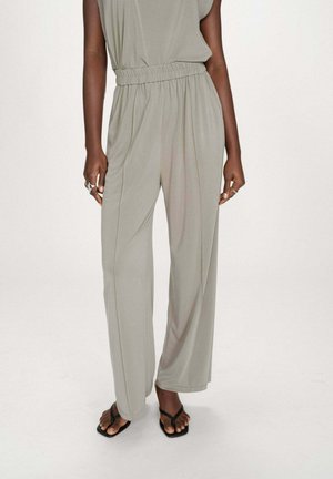 Femme portant un pantalon ample gris clair à taille élastique assorti de sandales noires à bout carré, debout devant un fond uni.