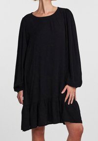 Vestido negro con cuello redondo, mangas largas con hombros fruncidos y un dobladillo acampanado con detalles de volantes. Hecho de una tela ligera.