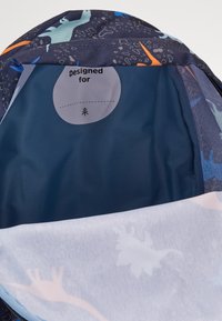 Mochila con patrón de dinosaurios muestra una etiqueta interior que dice "Diseñado para" con espacio para escribir un nombre.