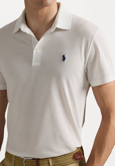 Camisa polo branca com um acabamento texturizado, apresentando uma pala de dois botões e um logótipo de cavalo azul-marinho no lado esquerdo do peito. Usada com um cinto castanho.