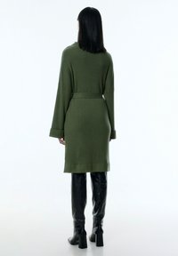 Olivgrünes Strickkleid mit tailliertem Gürtel, langen Ärmeln und gerippter Textur. Kombiniert mit schwarzen, knielangen Lederstiefeln.