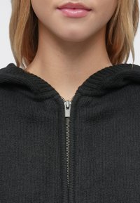 Μαύρο φούτερ με φερμουάρ και υφή υφάσματος, με μεταλλικό φερμουάρ και κουκούλα. Το υλικό φαίνεται μαλακό και ελαφρώς ριγωτό.