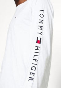 Maglietta a maniche lunghe bianca in cotone, con la scritta "TOMMY HILFIGER" in lettere nere lungo la manica sinistra e un piccolo logo sul petto.