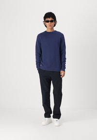 Marineblå strikkegenser med ribbestrikkede mansjetter og kant, kombinert med svarte bukser og hvite joggesko. Modell med mørke solbriller.