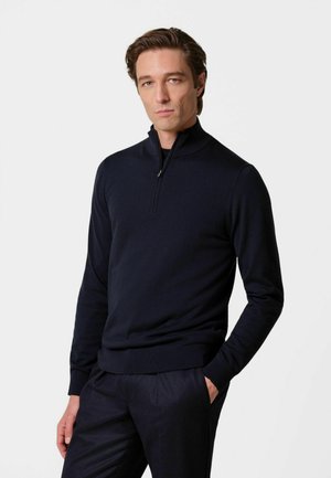Uomo che indossa un maglione nero aderente con zip a quarto e pantaloni neri, in piedi con una mano in tasca su uno sfondo bianco semplice.