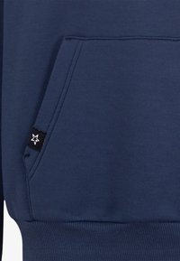 Mörkblå huvtröja med känguruficka, ribbstickade muddar och fåll. Inkluderar en liten svart etikett med en stjärnlogotyp på fickan.