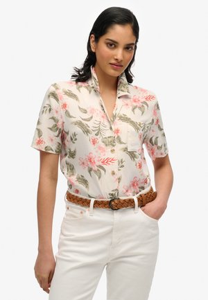 Femme portant une chemise blanche à manches courtes avec des motifs floraux roses et verts, rentrée dans un pantalon blanc avec une ceinture tressée marron.