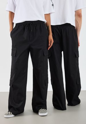 Schwarze Cargo-Hosen mit entspanntem Schnitt, ausgestattet mit Seitentaschen, einem elastischen Bund und einer glatten Stoffstruktur. Kombiniert mit karierten Slip-On-Schuhen.