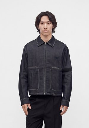 Jonge man met halflang donker haar die een donker spijkerjack met rits en contraststiksels en een zwarte broek draagt, staand voor een effen achtergrond.