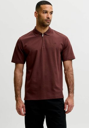 Homme portant un polo bordeaux à manches courtes avec une fermeture éclair, debout devant un fond gris clair uni.