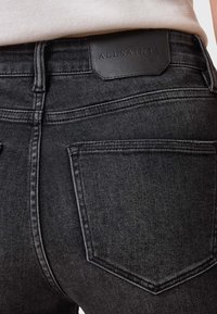 Jeans de mezclilla negra con un corte ajustado, que presentan costuras prominentes, bolsillos traseros y una etiqueta de cuero con texto en relieve en la cintura.