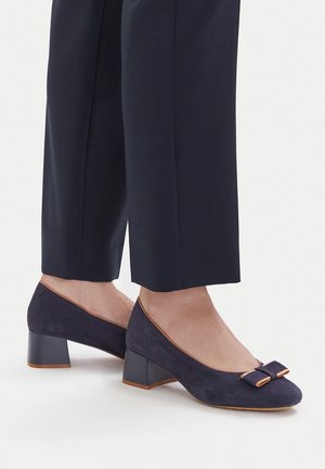 Marineblaue Wildleder-Schuhe mit niedrigem Blockabsatz und Schleifendetail, getragen mit schwarzen, verkürzten Hosen barfuß.