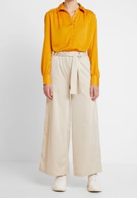 Žlutá hedvábná halenka s knoflíkovou légou a dlouhými rukávy, doplněná širokými béžovými kalhotami s vázáním v pase. Outfit je zakončen bílými teniskami.