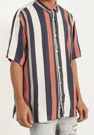 Homme portant une chemise à manches courtes à boutons avec des rayures verticales en bleu marine, blanc, rouille et beige, associée à un jean clair déchiré.