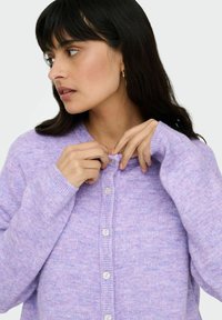 Cardigan en maille lilas à col rond avec une rangée de boutons sur le devant. Le tissu semble doux avec une finition légèrement texturée.